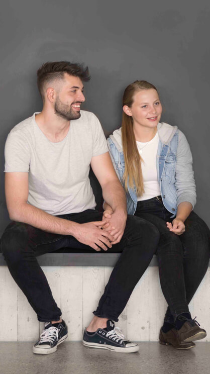 Eine junge Frau und ein junger Mann sitzen auf einer Bank und halten sich an den Haenden